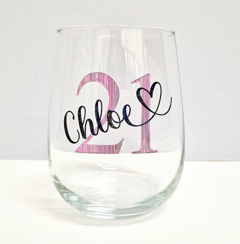 May include: A clear stemless wine glass with the number "21" in pink and a black heart. The name "Chloe" is written in cursive below the number. The glass is designed for a birthday celebration.