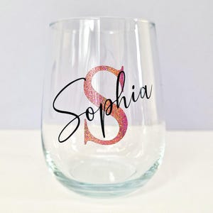 May include: A clear glass wine glass with a pink glitter initial "S" and the name "Sophia" written in black cursive.