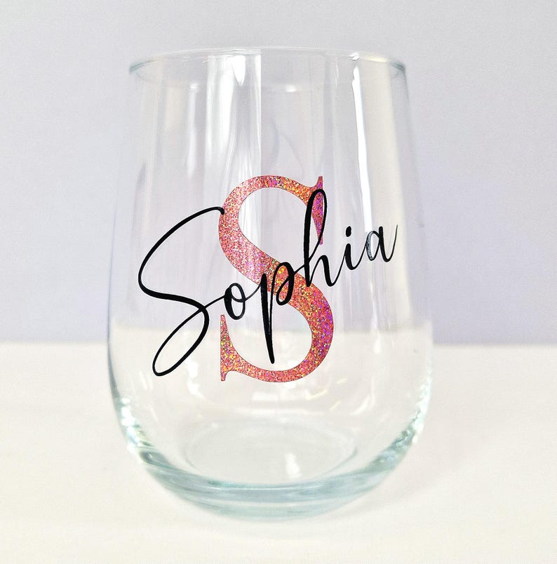 May include: A clear glass wine glass with a pink glitter initial "S" and the name "Sophia" written in black cursive.