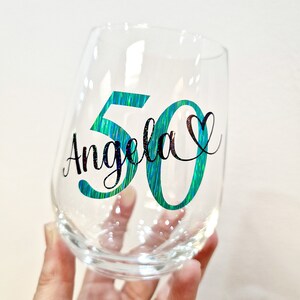 Op de afbeelding: Een helder steelloos wijnglas met de naam Angela in zwarte cursieve letters, geplaatst over een groot iriserend blauwgroen cijfer 50. Een klein zwart hartsymbool bevindt zich aan de rechterkant van de naam voor een decoratief effect.