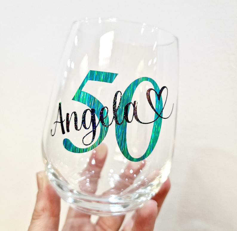 Op de afbeelding: Een helder steelloos wijnglas met de naam Angela in zwarte cursieve letters, geplaatst over een groot iriserend blauwgroen cijfer 50. Een klein zwart hartsymbool bevindt zich aan de rechterkant van de naam voor een decoratief effect.