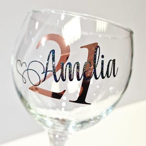 May include: A clear wine glass with a stem, featuring the number "21" in rose gold and the name "Amelia" in black script with glitter accents. A heart-shaped design is next to the name. The glass is designed for a birthday celebration.