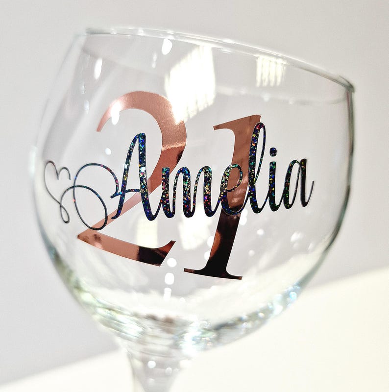 May include: A clear wine glass with a stem, featuring the number "21" in rose gold and the name "Amelia" in black script with glitter accents. A heart-shaped design is next to the name. The glass is designed for a birthday celebration.