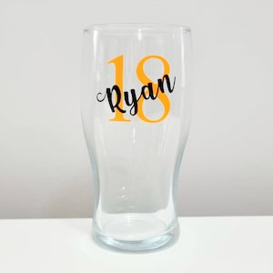 Puede incluir: Un vaso de pinta de vidrio transparente con una pegatina de vinilo negra y naranja que dice "18 Ryan".