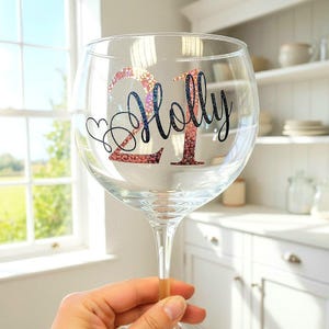 May include: A clear gin glass with the number "21" and the name "Holly" in a decorative font. The "21" is in a glittery pink and the name is in black script. A heart is drawn next to the name.