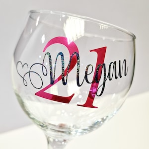 May include: A clear wine glass with a pink "21" and the name "Megan" in black script. The number "21" has a pink metallic finish. The word "Megan" has a glittery, iridescent finish. A heart design is next to the name.