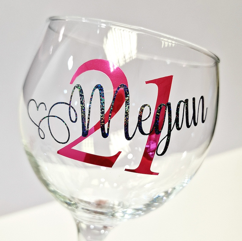 May include: A clear wine glass with a pink "21" and the name "Megan" in black script. The number "21" has a pink metallic finish. The word "Megan" has a glittery, iridescent finish. A heart design is next to the name.