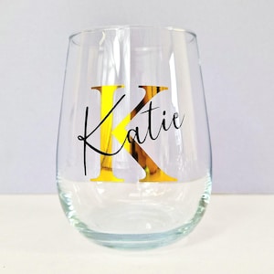 May include: A clear glass wine glass with a gold letter "K" and the name "Katie" in black lettering.