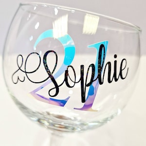 May include: A clear glass goblet with a stem, featuring the number "21" in iridescent blue and the name "Sophie" in black script. The glass has a rounded bowl shape and is decorated with small, scattered glitter accents.