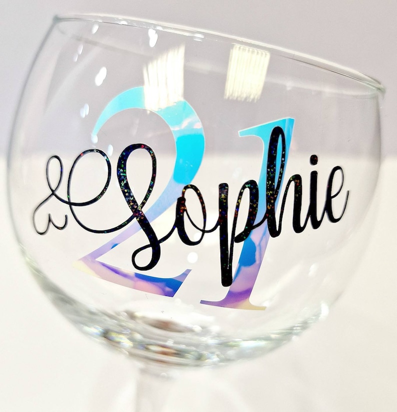 May include: A clear glass goblet with a stem, featuring the number "21" in iridescent blue and the name "Sophie" in black script. The glass has a rounded bowl shape and is decorated with small, scattered glitter accents.