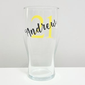 Puede incluir: Un vaso de pinta de vidrio transparente con una pegatina de vinilo amarilla que dice "Andrew 21" en letras negras.