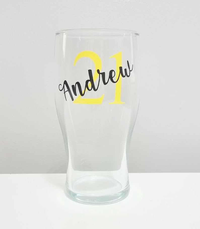 Puede incluir: Un vaso de pinta de vidrio transparente con una pegatina de vinilo amarilla que dice "Andrew 21" en letras negras.