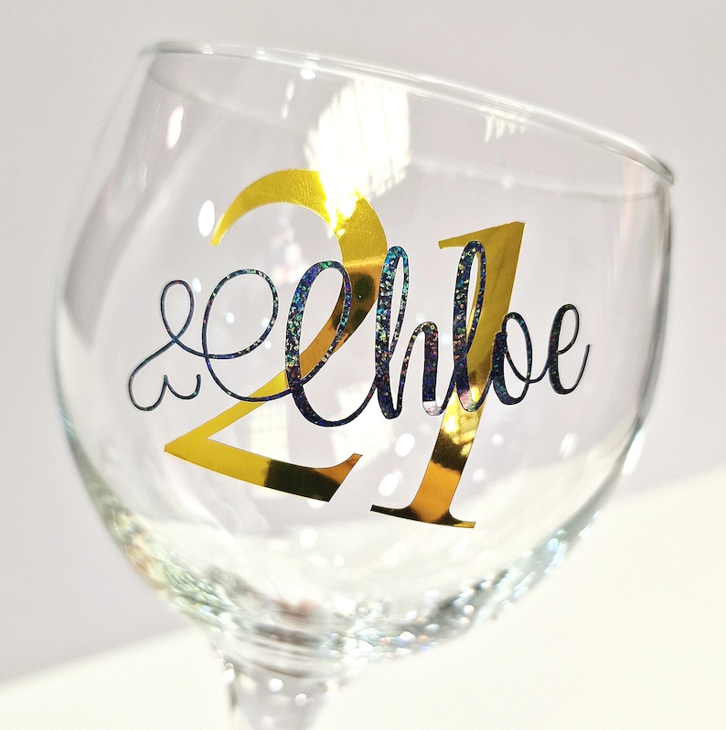 May include: A clear wine glass with a gold foil "21" and the name "Chloe" in a cursive font. The "Chloe" text has a shimmering, iridescent effect. The glass is tilted slightly.