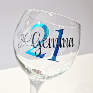 May include: A clear wine glass with a blue and glittery "21" and the name "Gemma" in black script. The glass is tilted, showcasing the design on the front. The background is a soft white.