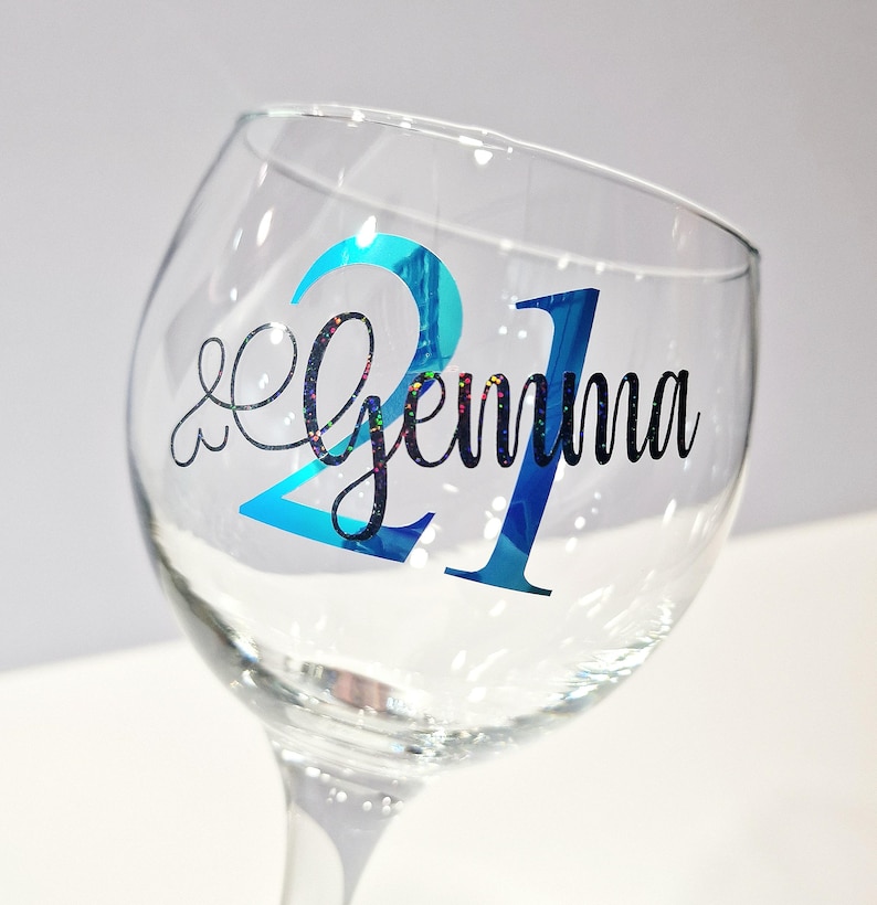 May include: A clear wine glass with a blue and glittery "21" and the name "Gemma" in black script. The glass is tilted, showcasing the design on the front. The background is a soft white.