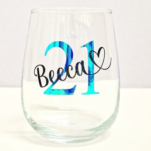 May include: A clear stemless wine glass with the number "21" in turquoise and the name "Becca" in black script. A black heart is next to the name. The glass is empty and sits on a white surface.