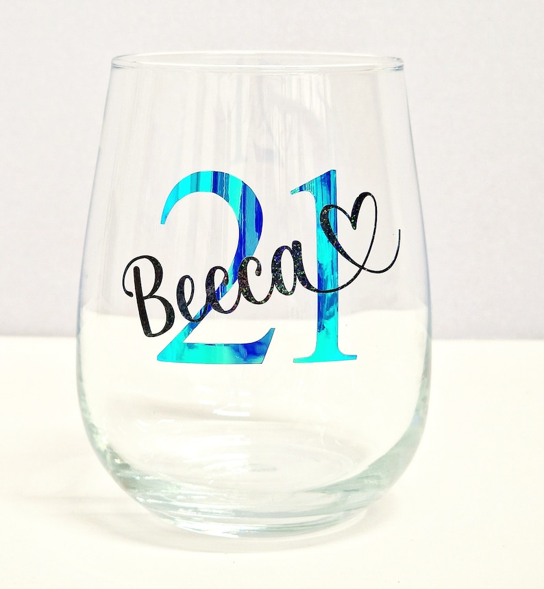 May include: A clear stemless wine glass with the number "21" in turquoise and the name "Becca" in black script. A black heart is next to the name. The glass is empty and sits on a white surface.