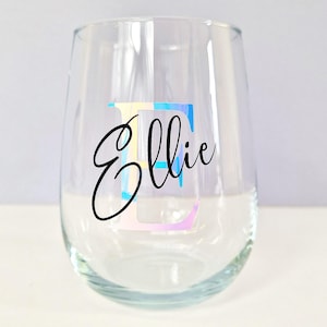 May include: A clear glass wine glass with the name "Ellie" written in black cursive script. The letter "E" is a large, colorful, iridescent sticker.