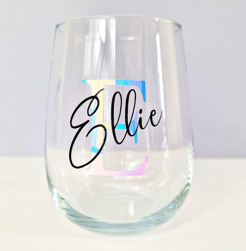 May include: A clear glass wine glass with the name "Ellie" written in black cursive script. The letter "E" is a large, colorful, iridescent sticker.