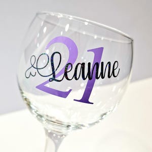 May include: A clear wine glass with a purple "21" and the name "Leanne" in black script. The "21" is a solid purple color, while the name has a glittery, iridescent effect. The glass is tilted slightly.