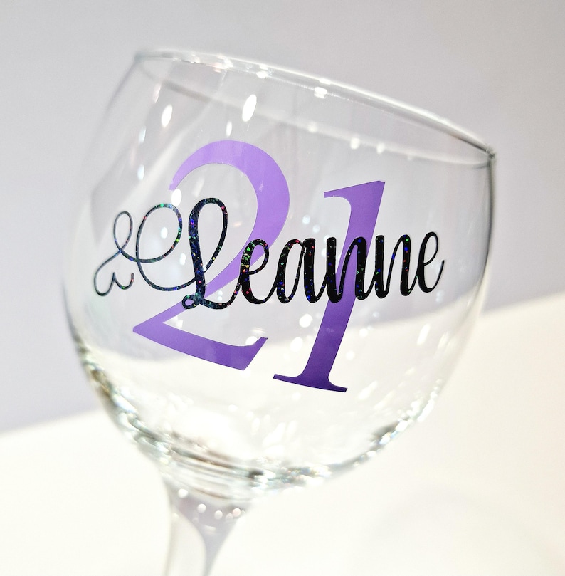 May include: A clear wine glass with a purple "21" and the name "Leanne" in black script. The "21" is a solid purple color, while the name has a glittery, iridescent effect. The glass is tilted slightly.
