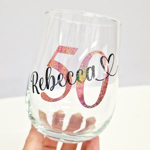 Op de afbeelding: Een helder, steelloos wijnglas met de naam Rebecca in zwarte cursieve letters, geplaatst over een groot glitterend roze getal 50. Een klein zwart hartsymbool staat aan het einde van de naam op dit gepersonaliseerde drinkglas.
