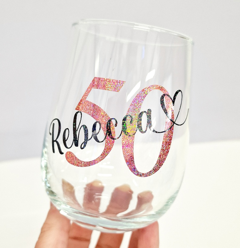 Op de afbeelding: Een helder, steelloos wijnglas met de naam Rebecca in zwarte cursieve letters, geplaatst over een groot glitterend roze getal 50. Een klein zwart hartsymbool staat aan het einde van de naam op dit gepersonaliseerde drinkglas.