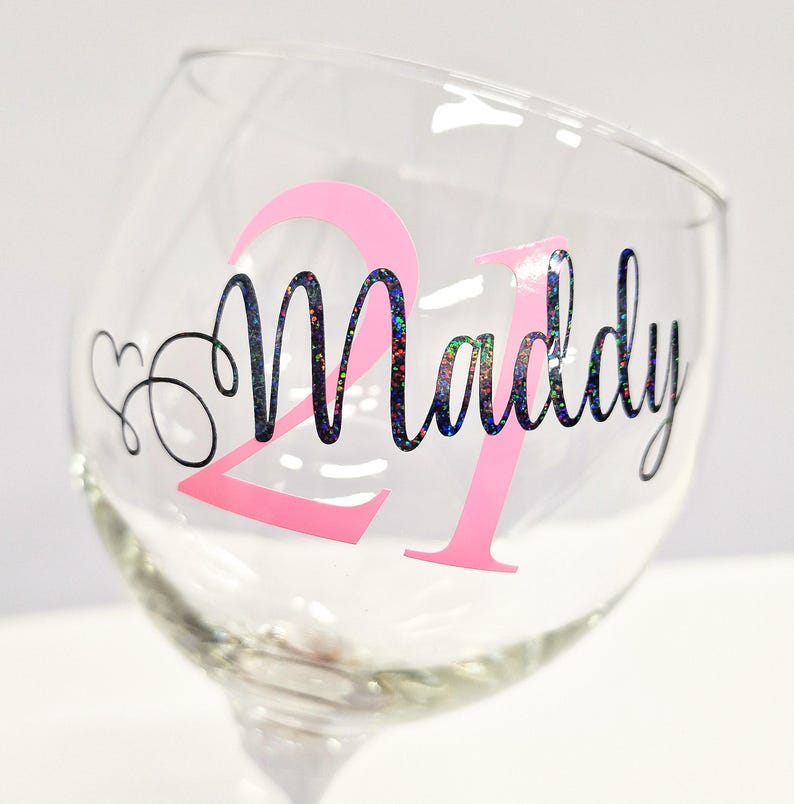 May include: A clear wine glass with a pink "21" and the name "Maddy" in a glittery font. A heart and swirls are next to the number 21. The glass is on a white background.