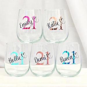 May include: Six clear stemless wine glasses, each personalized with a name and the number 21. The names are Emily, Katie, Hollie, Demi, and Beeca, each with a heart symbol. The numbers and names are in different colors.