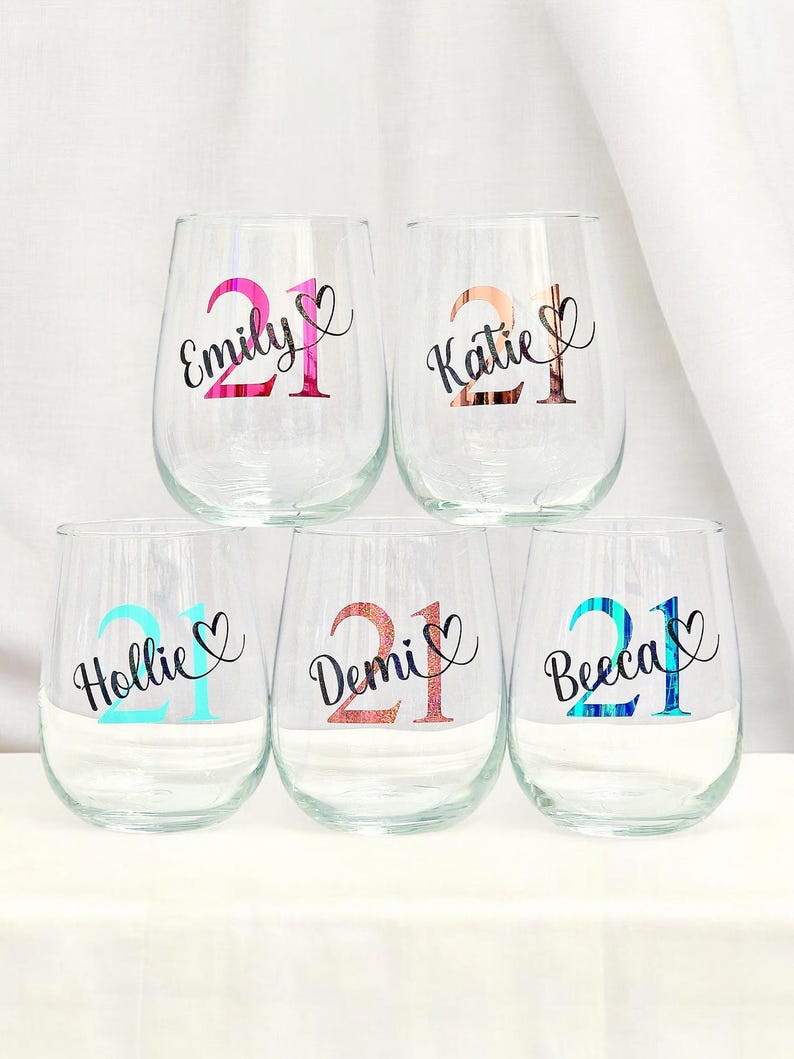 May include: Six clear stemless wine glasses, each personalized with a name and the number 21. The names are Emily, Katie, Hollie, Demi, and Beeca, each with a heart symbol. The numbers and names are in different colors.