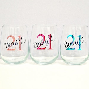 May include: Three stemless wine glasses with names and the number 21 in various colours. The names Demi, Emily, and Becca are in cursive script. The number 21 is in a glittery rose gold, pink, and teal colour.