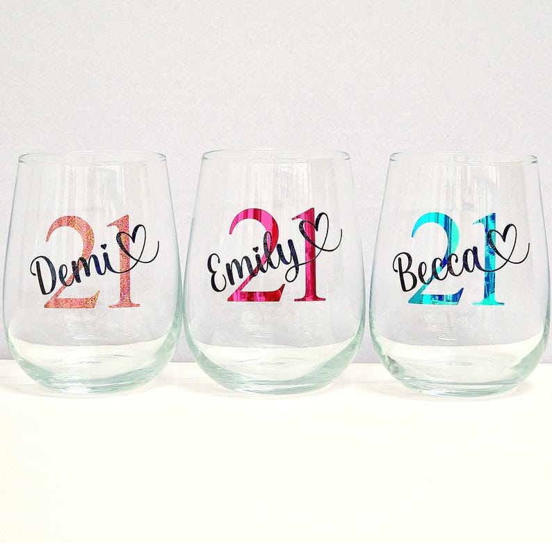 May include: Three stemless wine glasses with names and the number 21 in various colours. The names Demi, Emily, and Becca are in cursive script. The number 21 is in a glittery rose gold, pink, and teal colour.