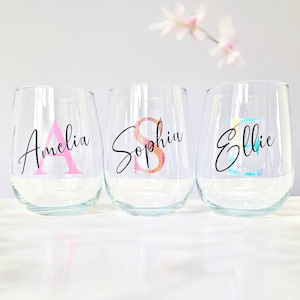 May include: Three clear glass wine glasses with personalized names and initial letters in pink, orange, and blue glitter. The names are Amelia, Sophia, and Ellie.