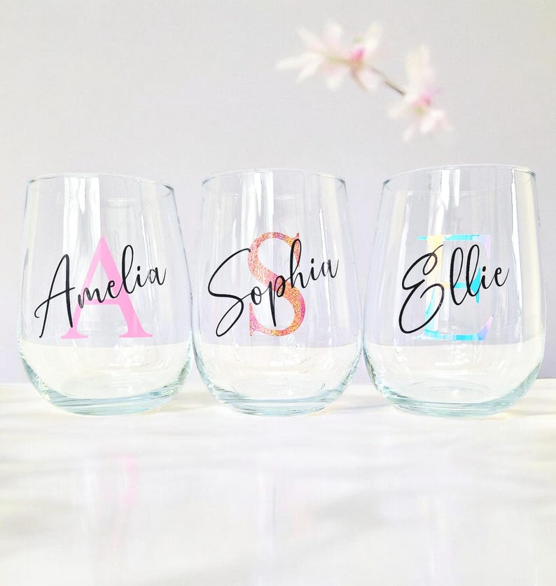 May include: Three clear glass wine glasses with personalized names and initial letters in pink, orange, and blue glitter. The names are Amelia, Sophia, and Ellie.