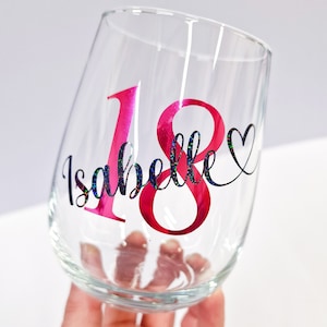 May include: A clear stemless wine glass with the name "Isabelle" and the number "18" in pink and black glitter lettering. A small heart is next to the number 8. The glass is held by a hand.