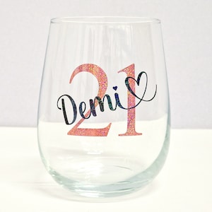 May include: A clear stemless wine glass featuring the number "21" in pink glitter and the name "Demi" in black script. A black heart is next to the number. The glass is empty and rests on a white surface.