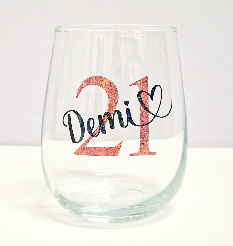 May include: A clear stemless wine glass featuring the number "21" in pink glitter and the name "Demi" in black script. A black heart is next to the number. The glass is empty and rests on a white surface.