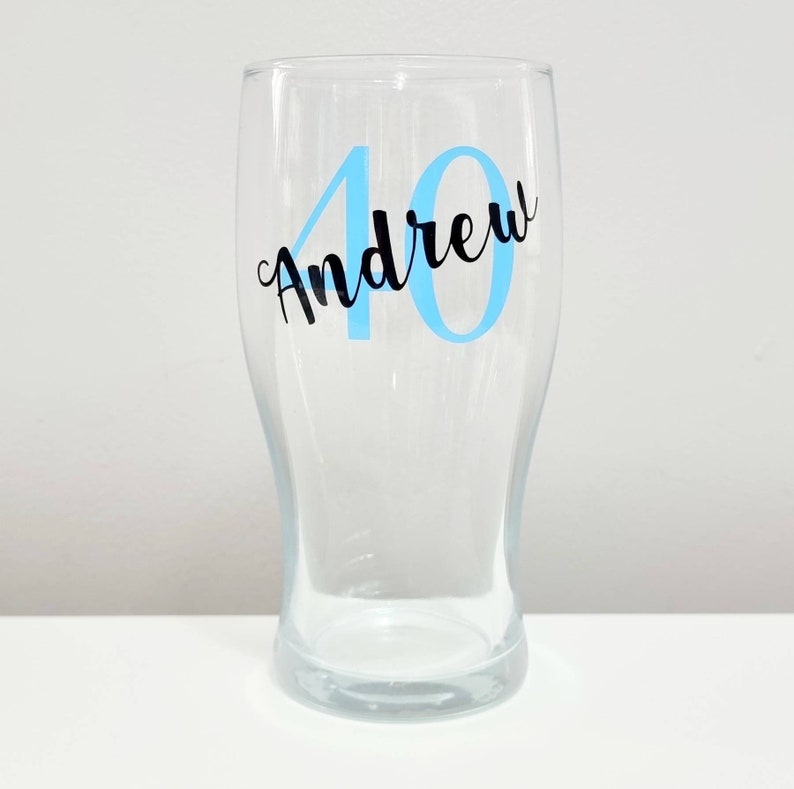 Puede incluir: Un vaso de pinta de vidrio transparente con una calcoman&iacute;a de vinilo azul y negro que dice "40 Andrew".
