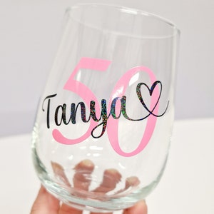 Op de afbeelding: Een helder wijnglas zonder steel met de naam Tanya in zwarte holografische letters, vergezeld van een groot roze cijfer 50 en een kleine zwarte hartomtrek. Het ontwerp is aangebracht op het buitenoppervlak van het glas.