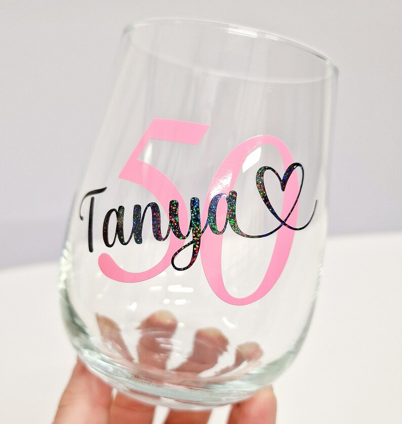 Op de afbeelding: Een helder wijnglas zonder steel met de naam Tanya in zwarte holografische letters, vergezeld van een groot roze cijfer 50 en een kleine zwarte hartomtrek. Het ontwerp is aangebracht op het buitenoppervlak van het glas.