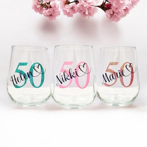Op de afbeelding: Drie doorzichtige wijnglazen zonder steel met gepersonaliseerde namen en het getal 50. Het eerste glas toont de naam Helen in zwarte letters over een turquoise 50, het tweede Nikki over een roze 50 en het derde Marie over een glinsterende oranje 50.