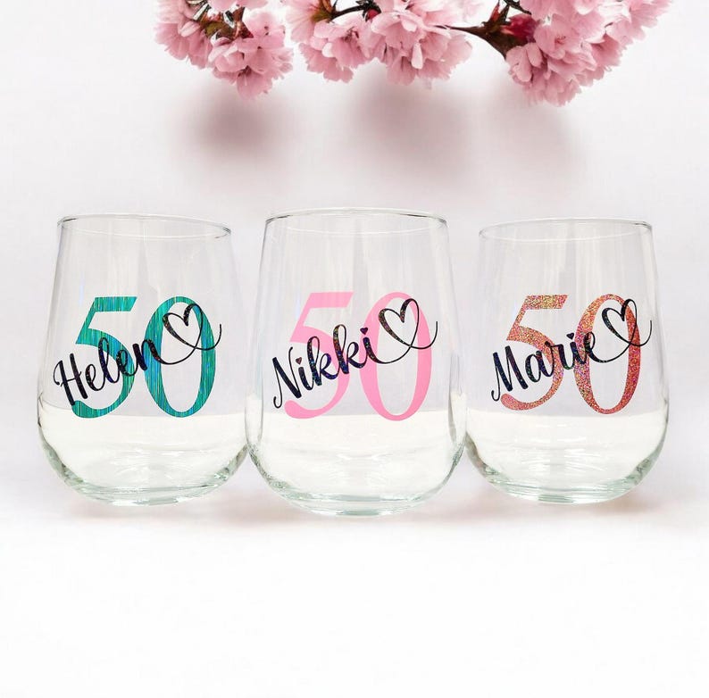 Op de afbeelding: Drie doorzichtige wijnglazen zonder steel met gepersonaliseerde namen en het getal 50. Het eerste glas toont de naam Helen in zwarte letters over een turquoise 50, het tweede Nikki over een roze 50 en het derde Marie over een glinsterende oranje 50.