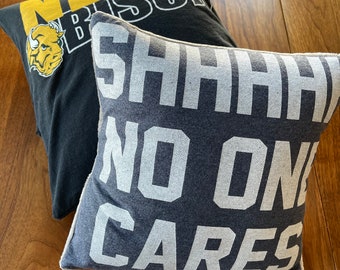 Puede incluir: Dos almohadas decorativas. Una almohada es negra con texto blanco que dice "NDSU Bison" y un logotipo de bisonte amarillo. La otra almohada es gris con texto blanco que dice "SHHH NO ONE CARES".