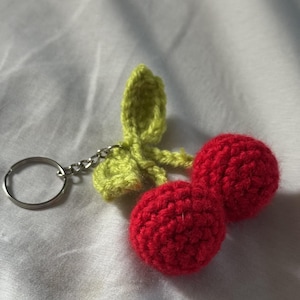 May include: A handmade crochet cherry keychain. The keychain features two red cherries with a green leaf and stem. The keychain is attached to a silver metal ring.