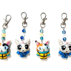 May include: Four keychains featuring sculpted cat figures with large eyes. Two cats are calico with yellow outfits, and two are silver with blue outfits and nurse caps. Each keychain has a silver lobster clasp and a strand of blue and yellow beads.