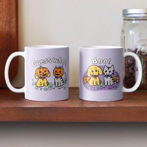 May include: Two white ceramic mugs with Halloween-themed illustrations. One mug says "Guess who?" and features two dogs with pumpkin heads. The other mug says "Boo!" and features a dog and a cat with pumpkin heads.