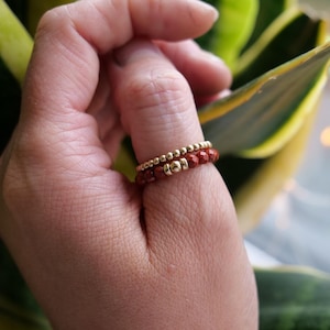 May include: Two gold rings with red beads on a finger. The rings are stacked on top of each other.