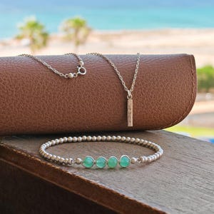 May include: A silver necklace with a rectangular pendant and a beaded bracelet featuring four green gemstones. The jewellery is displayed on a wooden surface with a brown leather case behind. The ocean and sky are visible in the background.