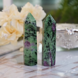 May include: Two green, black, and pink gemstone towers, also known as zoisite, on a white surface.