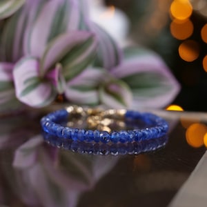 Puede incluir: Un brazalete de piedras preciosas azules con un cierre de oro. El brazalete está hecho de pequeñas cuentas redondas y está sobre una superficie reflectante.